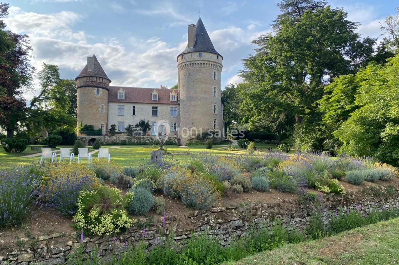 Château en pierre entouré de jardins fleuris, avec des arbres et un ciel partiellement nuageux en arrière-plan.