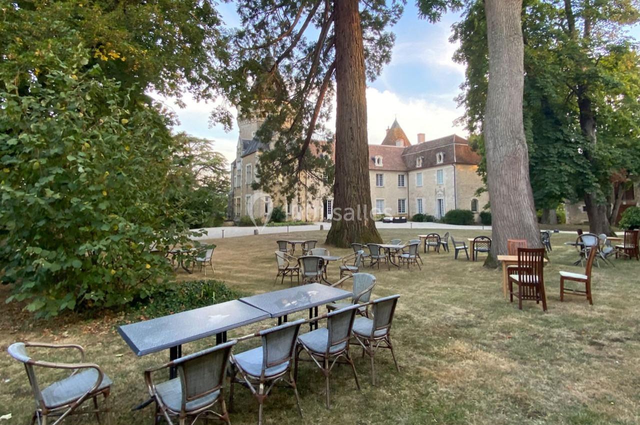 Tables et chaises disposées sur une pelouse ombragée devant un bâtiment historique entouré d'arbres.