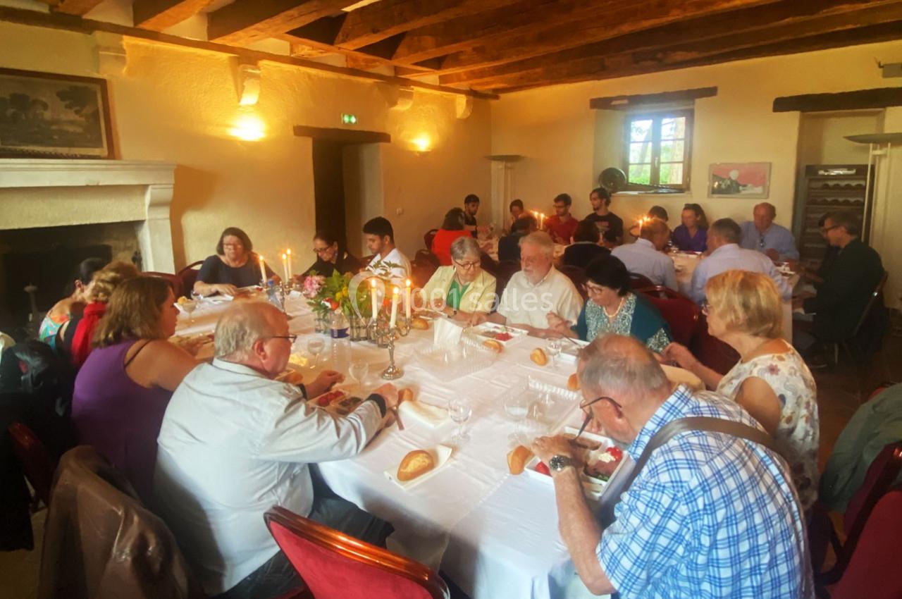Personnes d'âges variés partageant un repas autour d'une grande table dans une salle chaleureuse avec poutres apparentes.