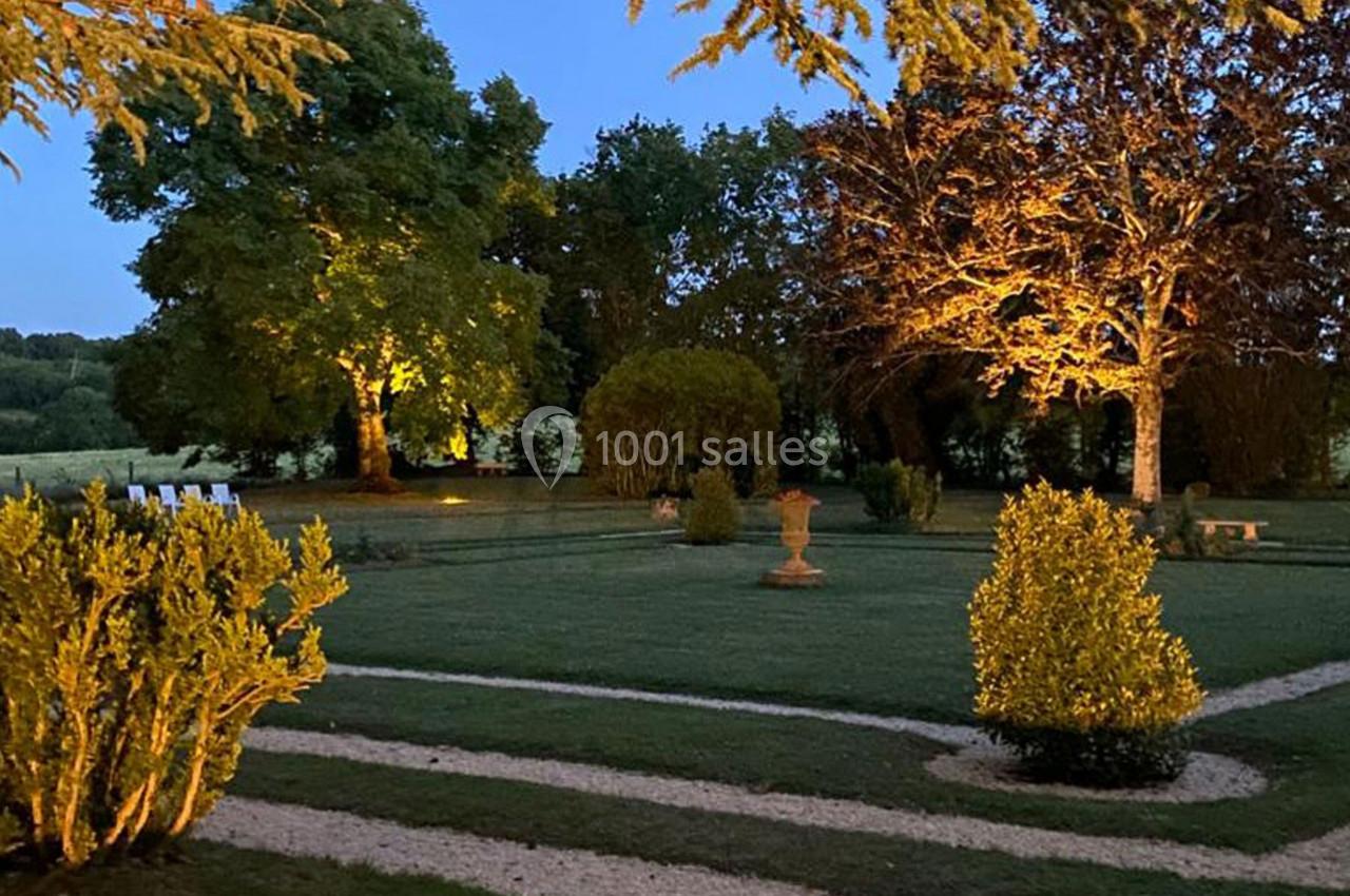 Jardin éclairé au crépuscule avec pelouse, allées gravillonnées, arbustes taillés et arbres illuminés.