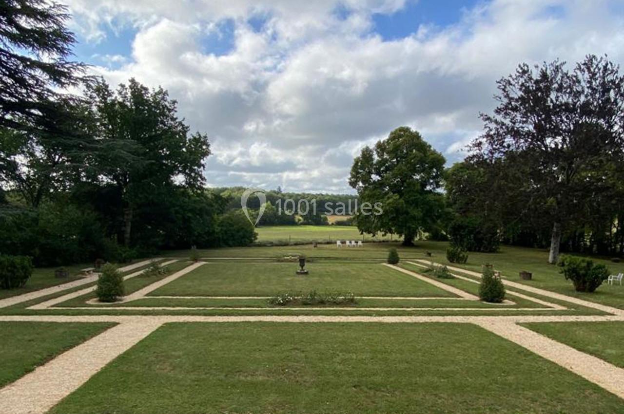 Vue d'un jardin symétrique avec pelouses, allées gravillonnées et arbres, sous un ciel partiellement nuageux.