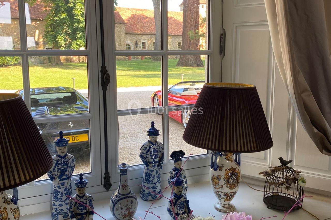 Vue depuis une fenêtre sur une cour avec deux voitures de sport, des lampes et des porcelaines décoratives au premier plan.