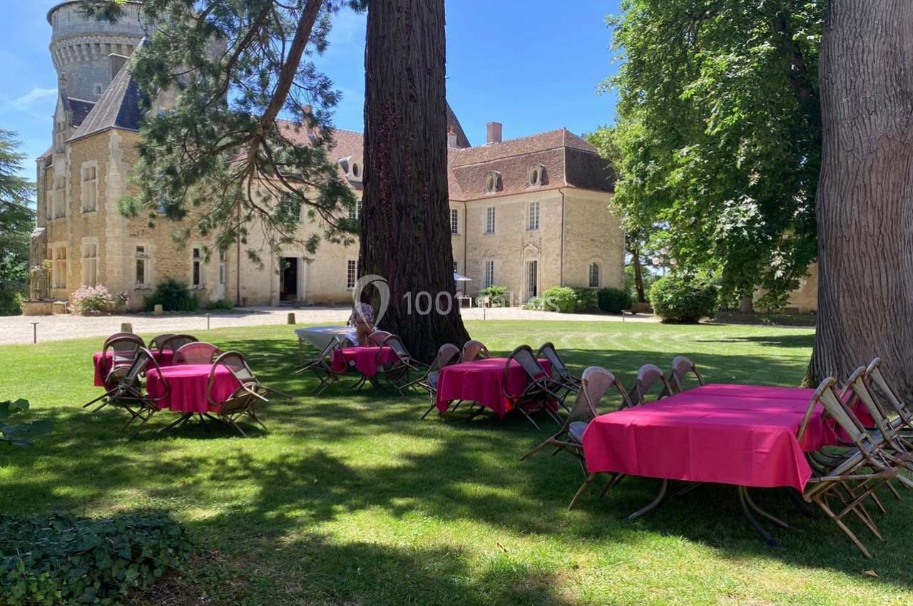 Tables et chaises avec nappes roses disposées sur une pelouse ombragée devant un bâtiment en pierre.