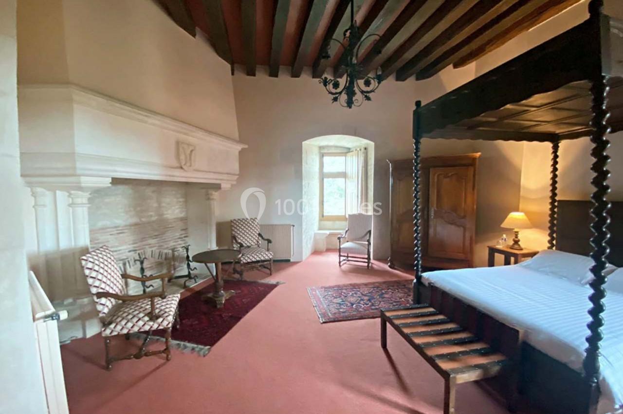 Chambre avec lit à baldaquin, cheminée en pierre, meubles anciens, tapis rouge et fenêtre donnant sur l'extérieur.