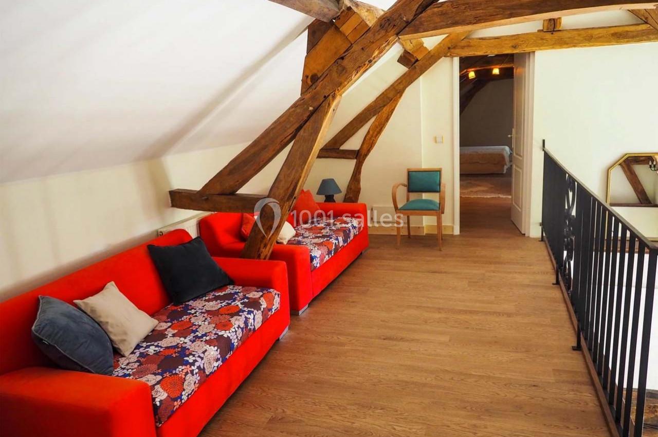 Salon mansardé avec poutres apparentes, deux canapés rouges, parquet en bois clair et chaise bleue près d'une rambarde.