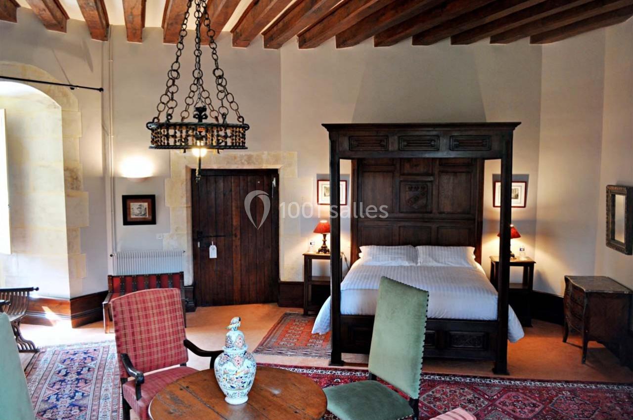 Chambre avec lit à baldaquin en bois, mobilier ancien, tapis colorés et plafond avec poutres apparentes.