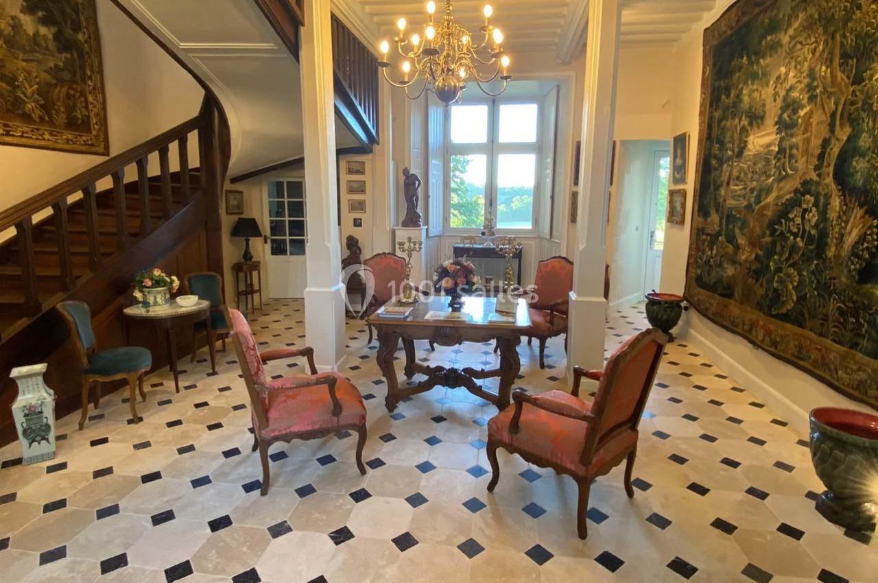 Salle élégante avec mobilier ancien, tapisseries murales, sol en damier et escalier en bois dans un intérieur lumineux.