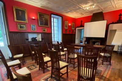Bâtiment en pierre avec toit en tuiles rouges, entouré d'arbres et d'une pelouse, sous un ciel partiellement nuageux.