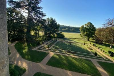 Bâtiment en pierre avec toit en tuiles rouges, entouré d'arbres et d'une pelouse, sous un ciel partiellement nuageux.