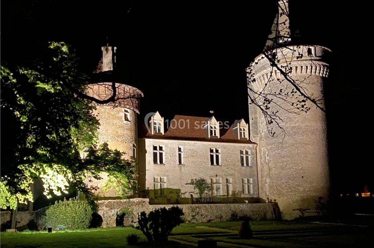 Château médiéval éclairé de nuit, entouré d'arbres et d'un jardin visible au premier plan.