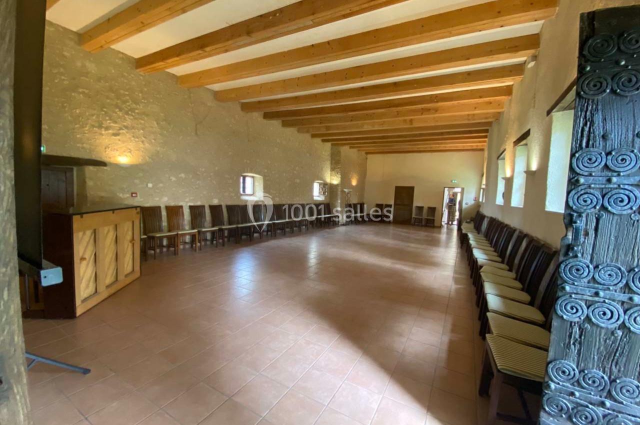 Salle spacieuse avec murs en pierre, poutres apparentes, chaises alignées et lumière naturelle entrant par des fenêtres.