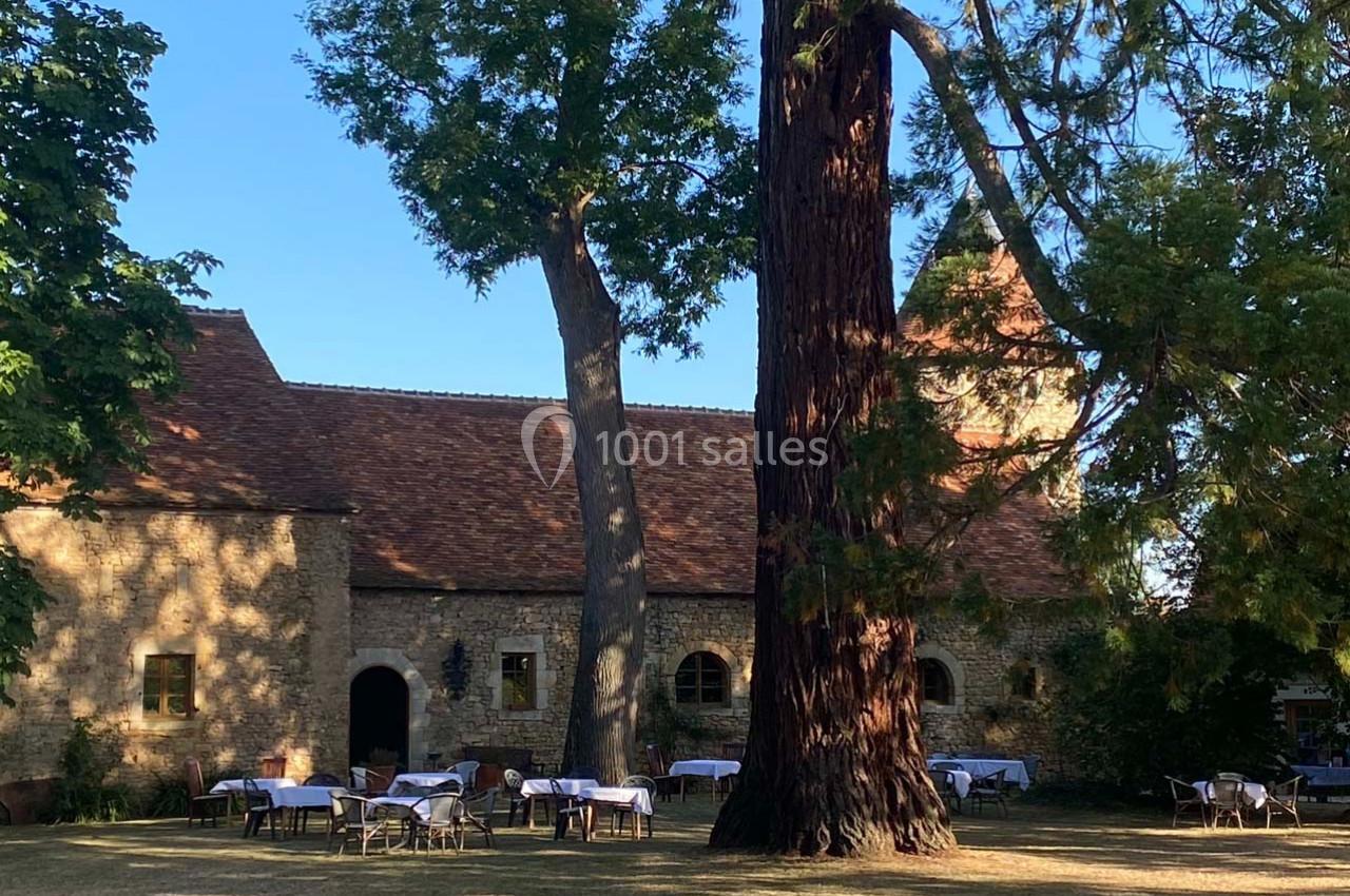 Cour extérieure d'un bâtiment en pierre avec des tables dressées sous de grands arbres, par une journée ensoleillée.