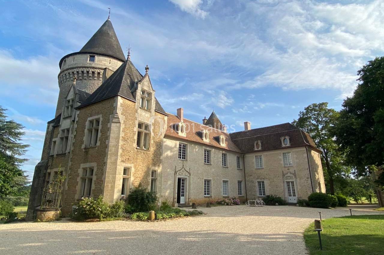 Château en pierre avec tour ronde, entouré d'arbres, sous un ciel partiellement nuageux.