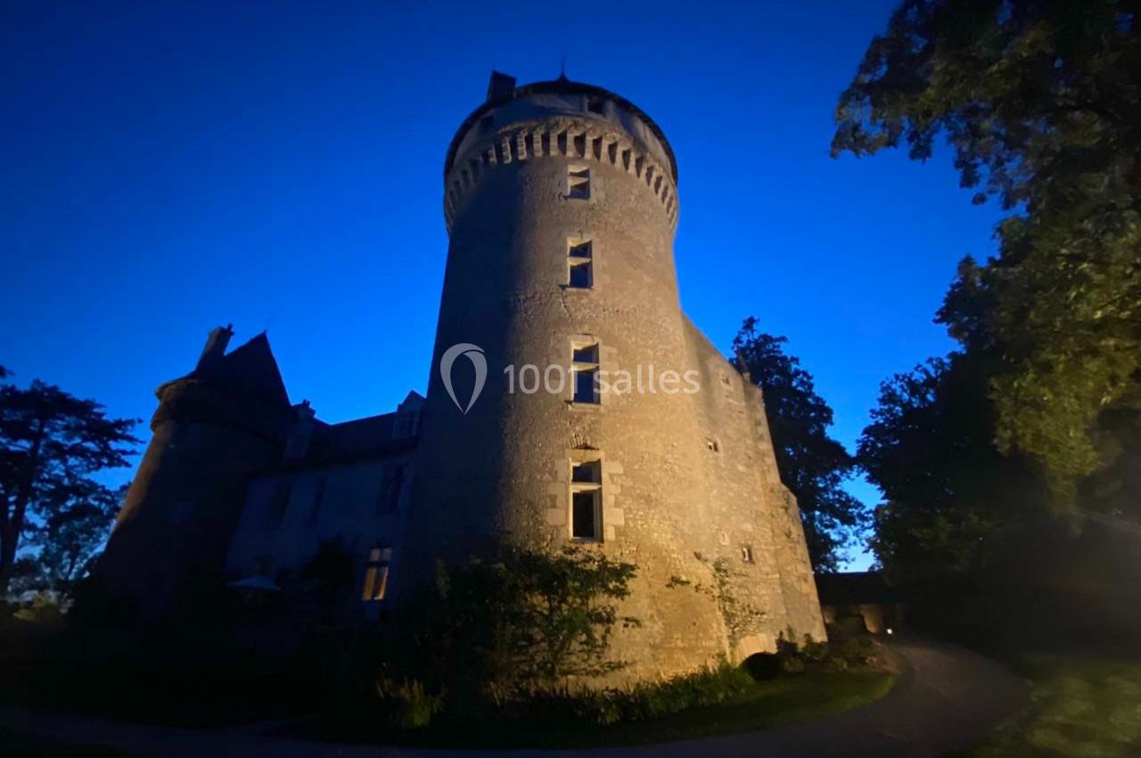 Château en pierre éclairé à la tombée de la nuit, entouré d'arbres et d'un chemin sinueux.