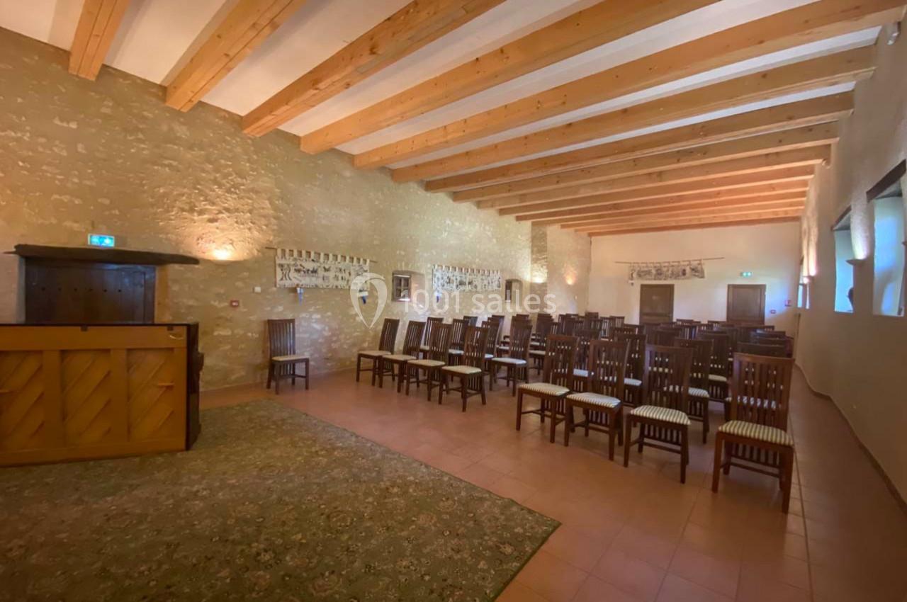 Salle avec des murs en pierre, plafond à poutres apparentes, chaises alignées et un piano en bois à gauche.