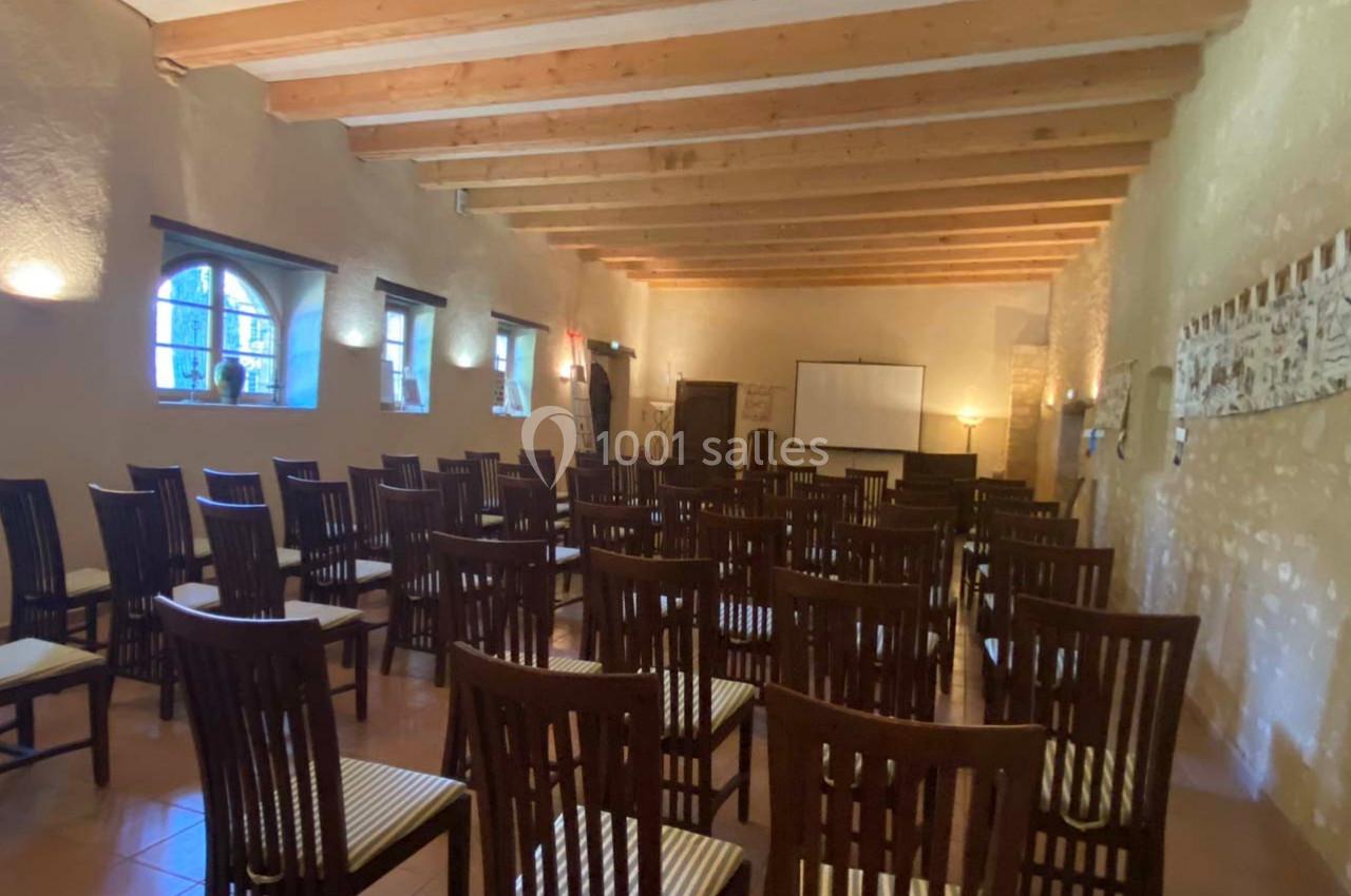 Salle avec des chaises en bois alignées, murs en pierre, poutres apparentes et un écran de projection au fond.