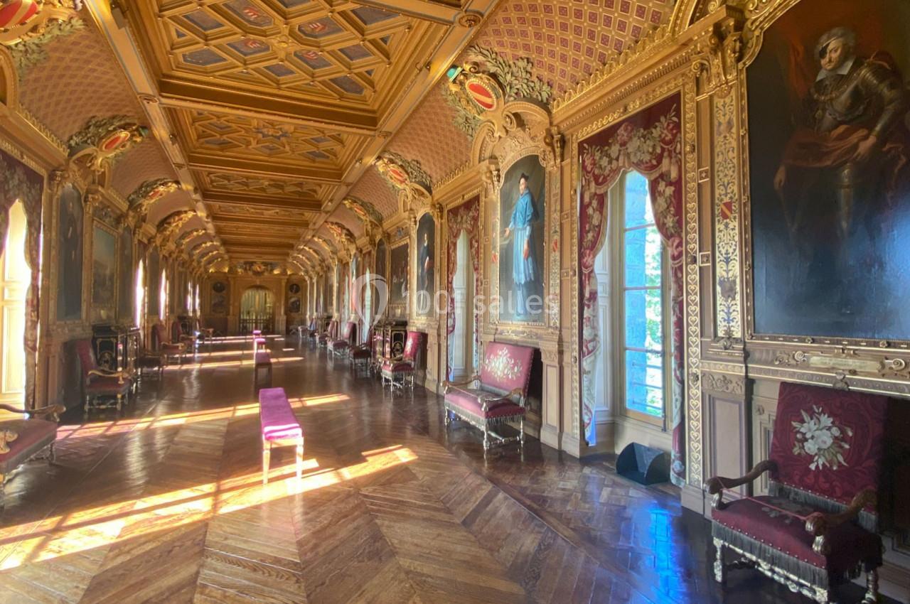 Galerie ornée de peintures et de dorures, avec parquet en bois et lumière naturelle entrant par des fenêtres.