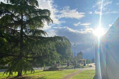 Bâtiment en pierre avec toit en tuiles rouges, entouré d'arbres et d'une pelouse, sous un ciel partiellement nuageux.