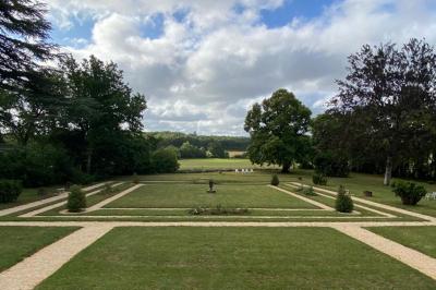 Bâtiment en pierre avec toit en tuiles rouges, entouré d'arbres et d'une pelouse, sous un ciel partiellement nuageux.