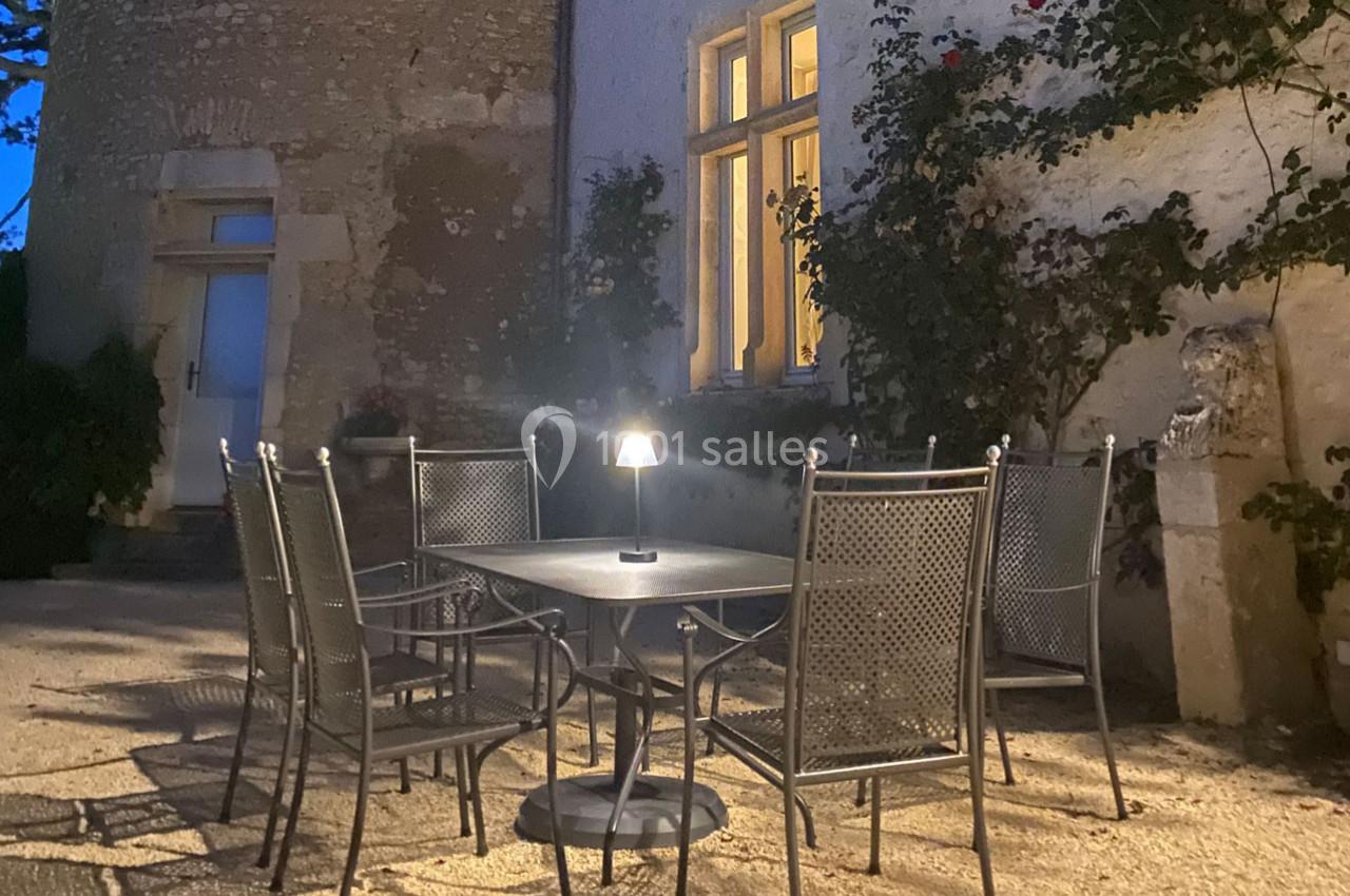 Table et chaises en métal sur une terrasse éclairée par une lampe, devant un mur en pierre avec des fenêtres et des rosiers.