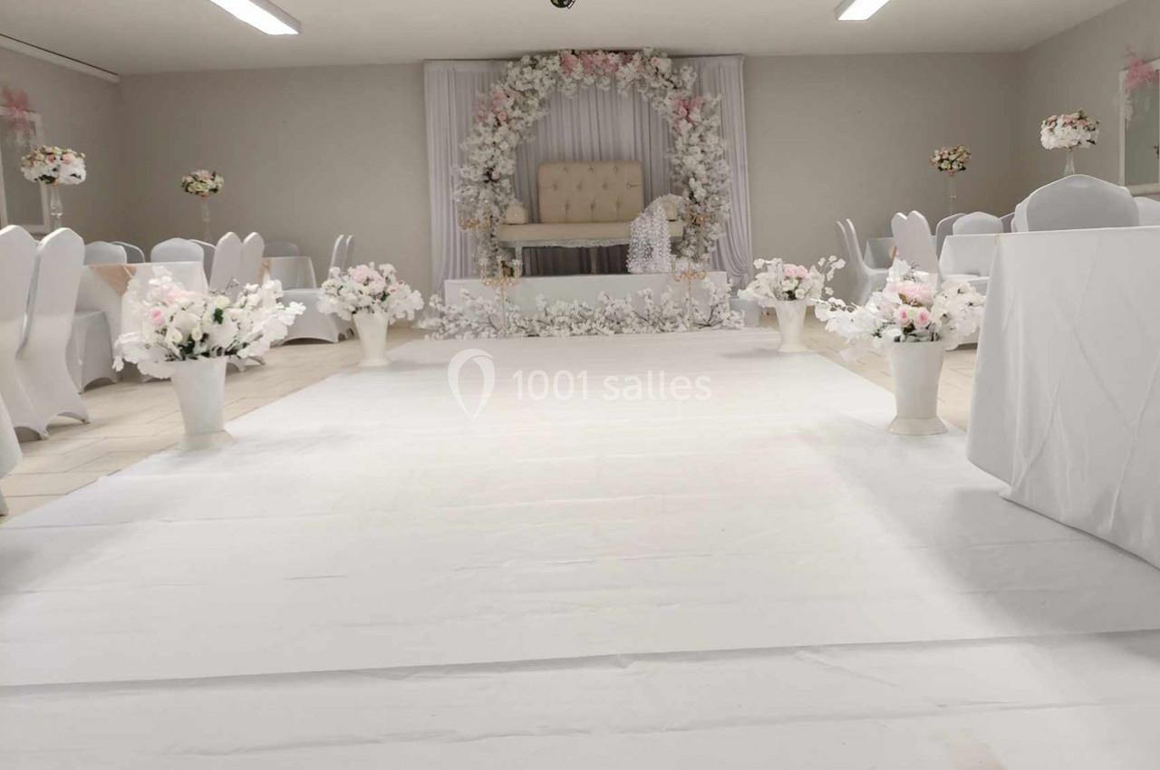 Salle de réception décorée en blanc avec des fleurs et une arche florale autour d'un fauteuil central.