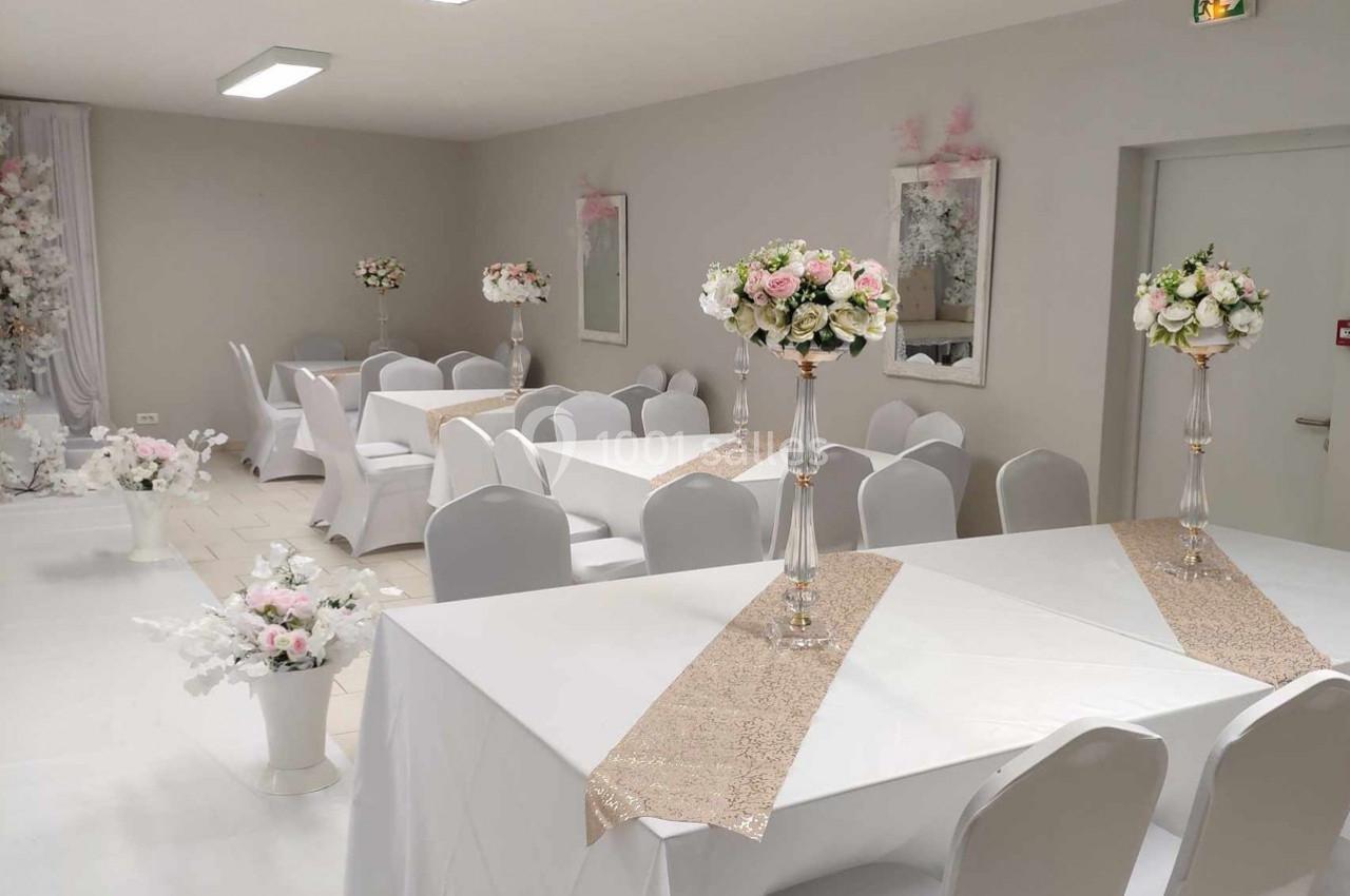 Salle de réception décorée avec des tables blanches, chemins de table beiges et centres de table fleuris.
