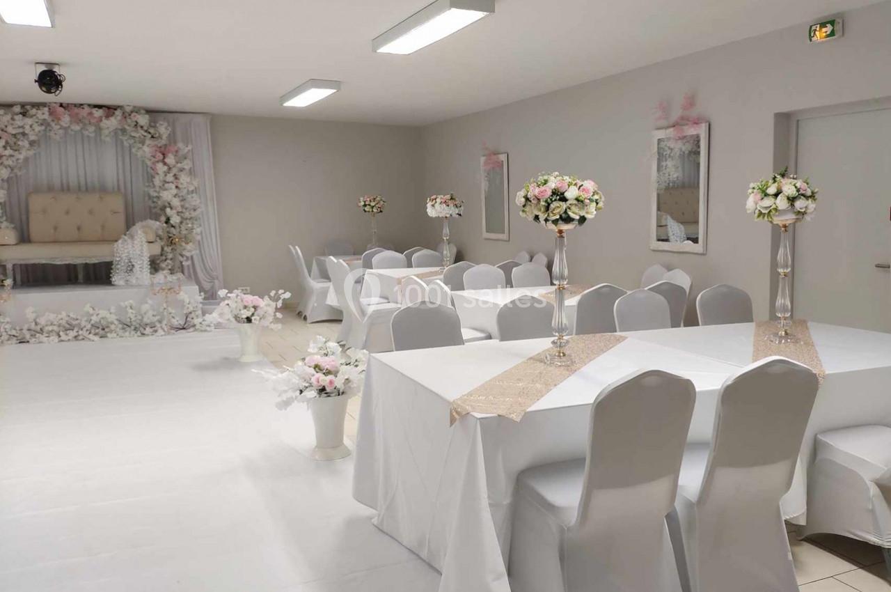 Salle de réception décorée avec des tables blanches, des chemins de table beiges et des compositions florales élégantes.