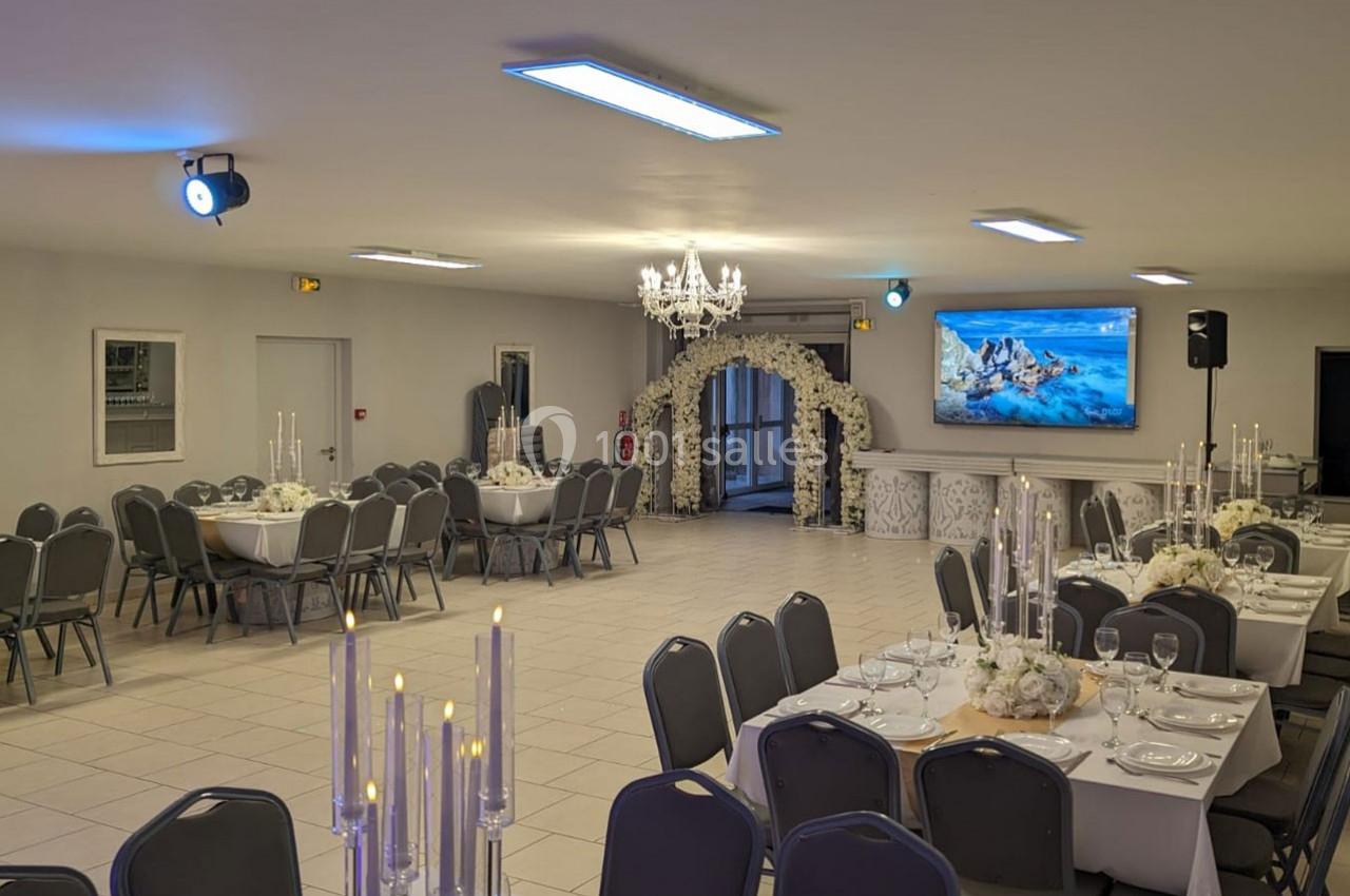 Salle de réception décorée avec des tables dressées, un lustre central, un écran mural et une arche fleurie.