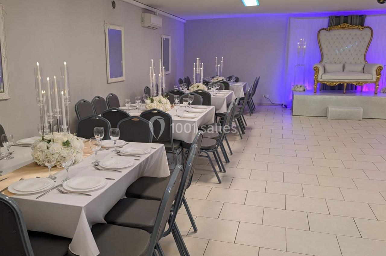 Salle de réception décorée avec des tables dressées, chandeliers et un fauteuil élégant sur une estrade.
