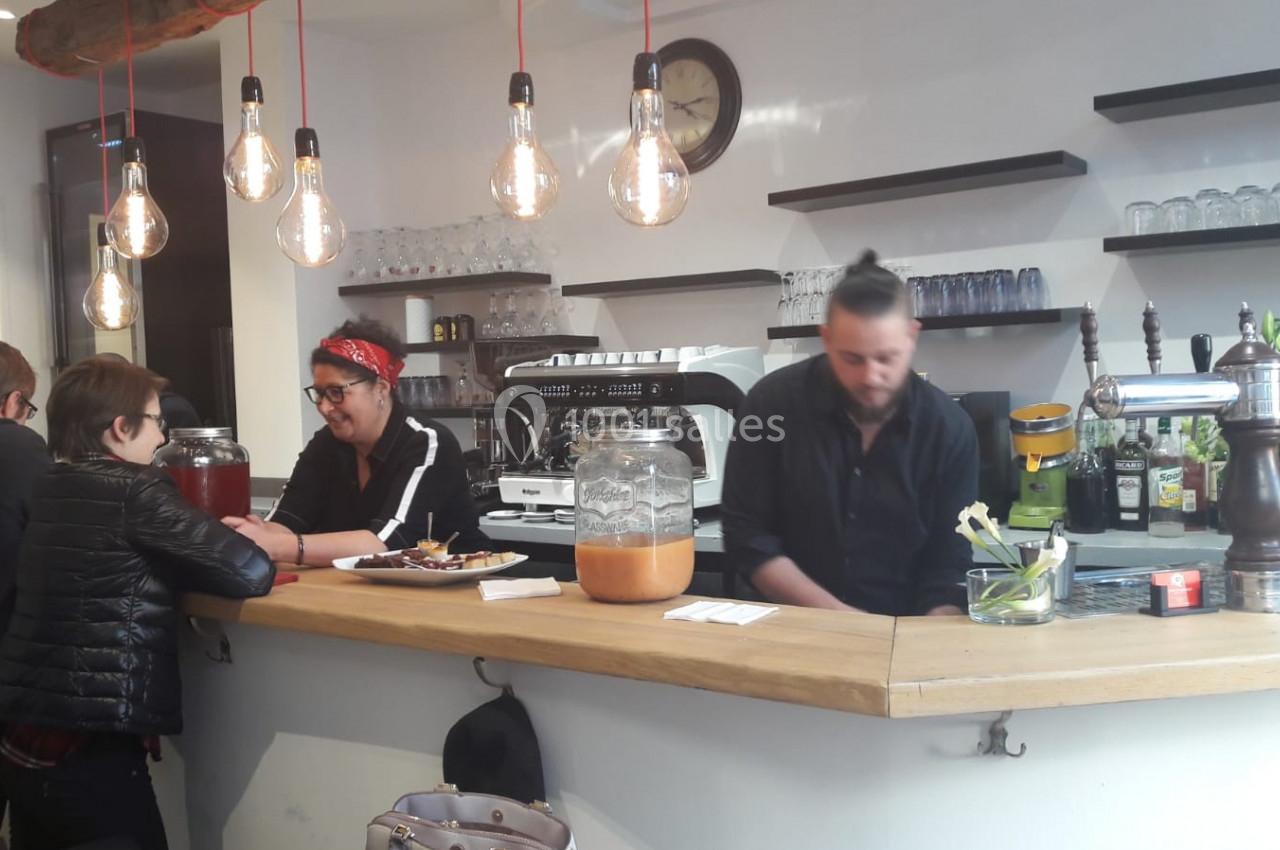 Deux personnes discutent au comptoir d'un café, avec un serveur préparant des boissons en arrière-plan.