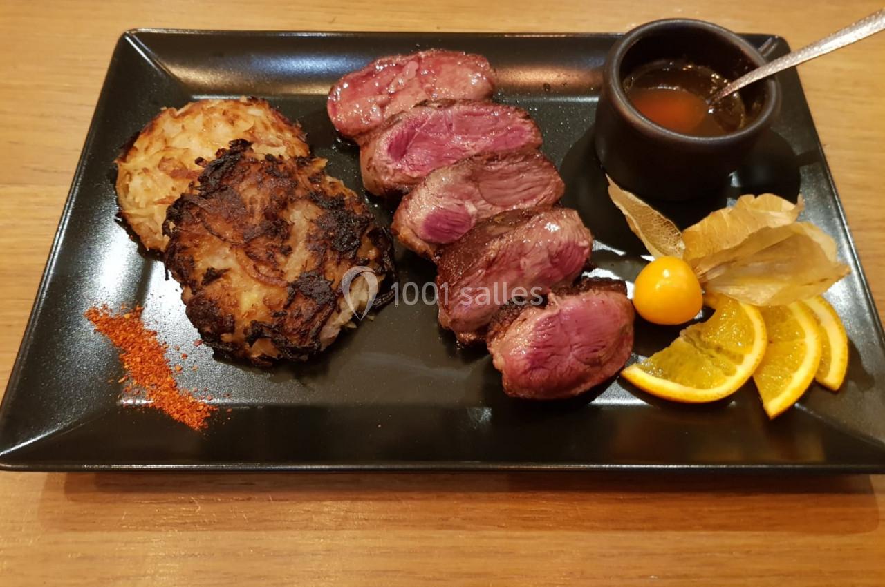 Assiette contenant des tranches de viande rosée, une galette de pommes de terre, une sauce et des garnitures d'agrumes.