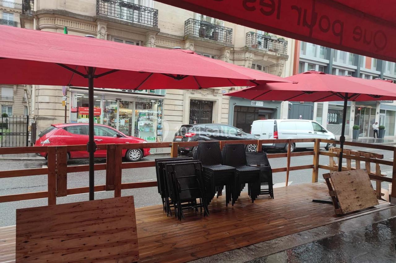 Terrasse en bois sous des parasols rouges, avec des chaises empilées, sur une rue bordée de bâtiments et de voitures.