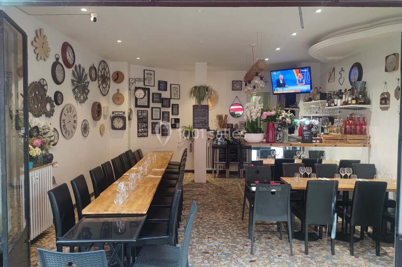 Salle de restaurant avec une longue table en bois, des chaises noires, des décorations murales et un bar au fond.