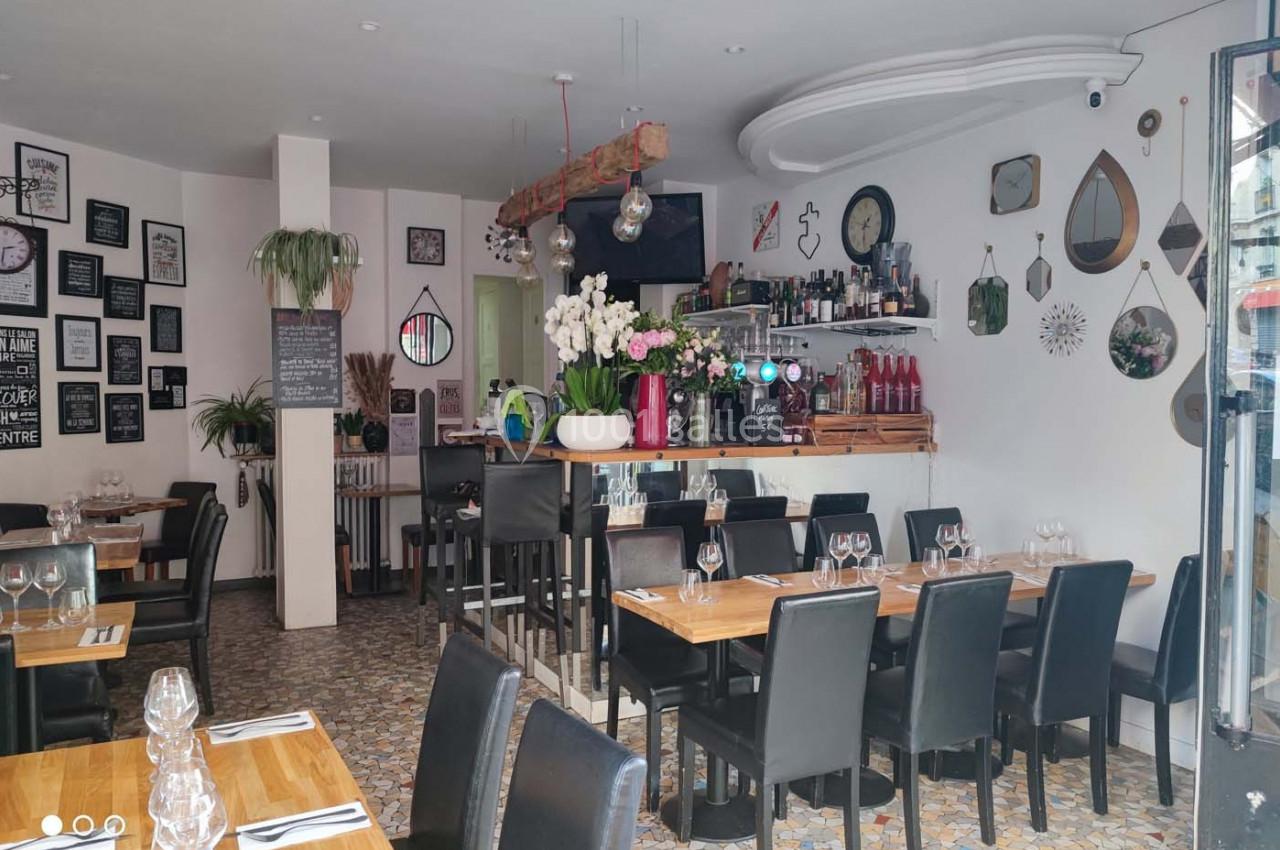 Salle de restaurant lumineuse avec tables en bois, chaises noires, décoration murale et bar en arrière-plan.