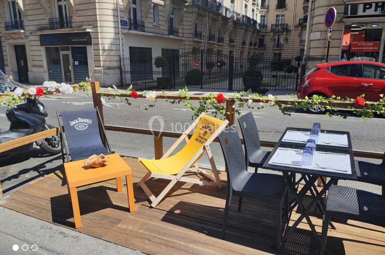 Terrasse en bois avec chaises, table dressée et fleurs, située en bord de rue devant des immeubles urbains.