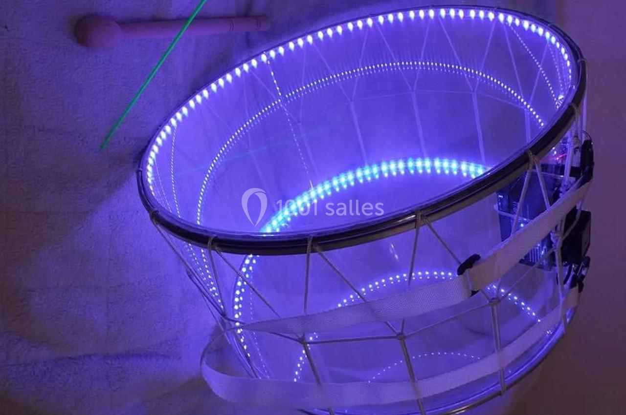 Tambour transparent éclairé par des LED violettes, posé sur une surface claire avec une baguette et une tige verte.