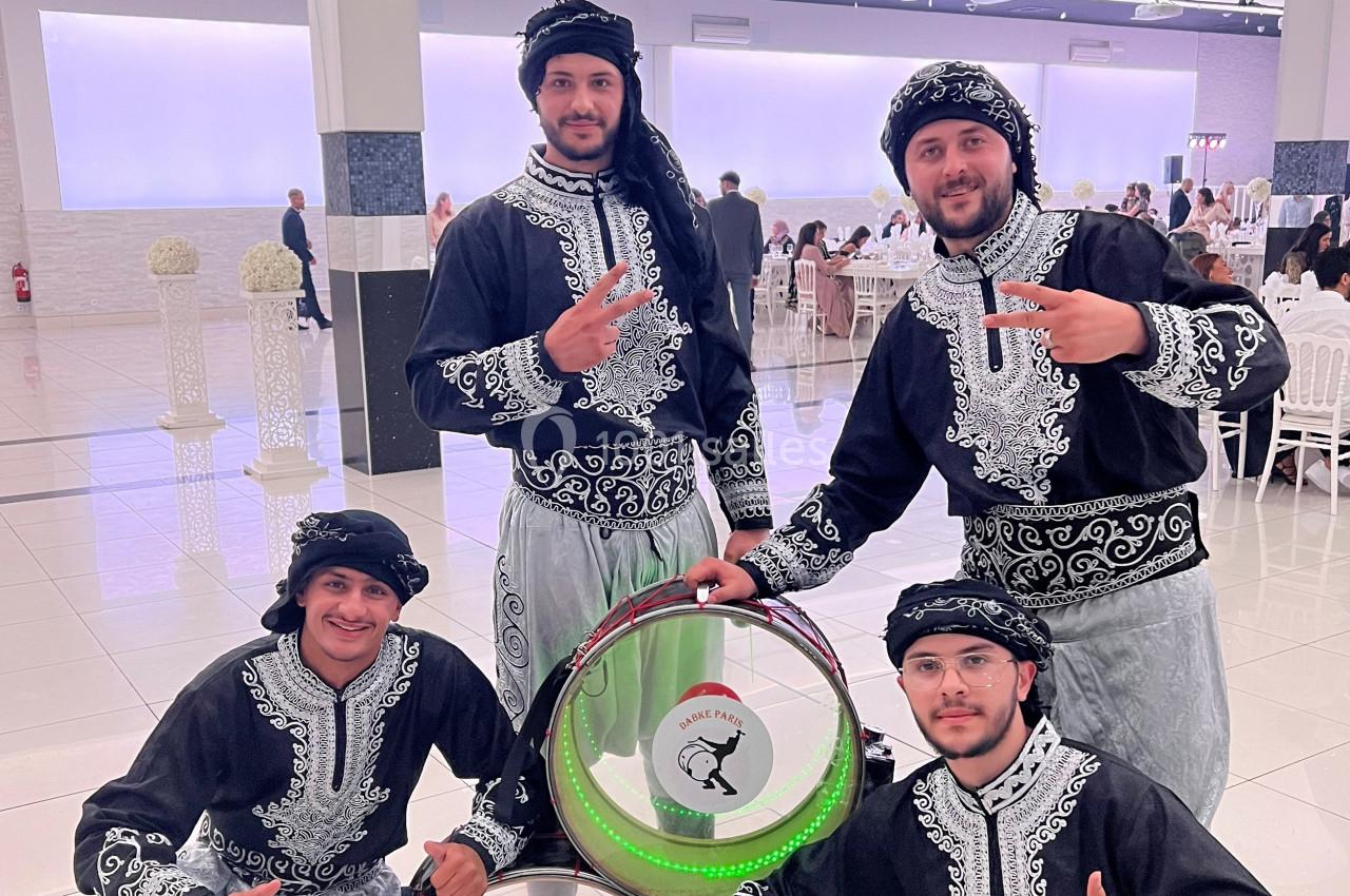 Quatre hommes en tenue traditionnelle posent autour d'un tambour décoré, dans une salle de réception lumineuse.