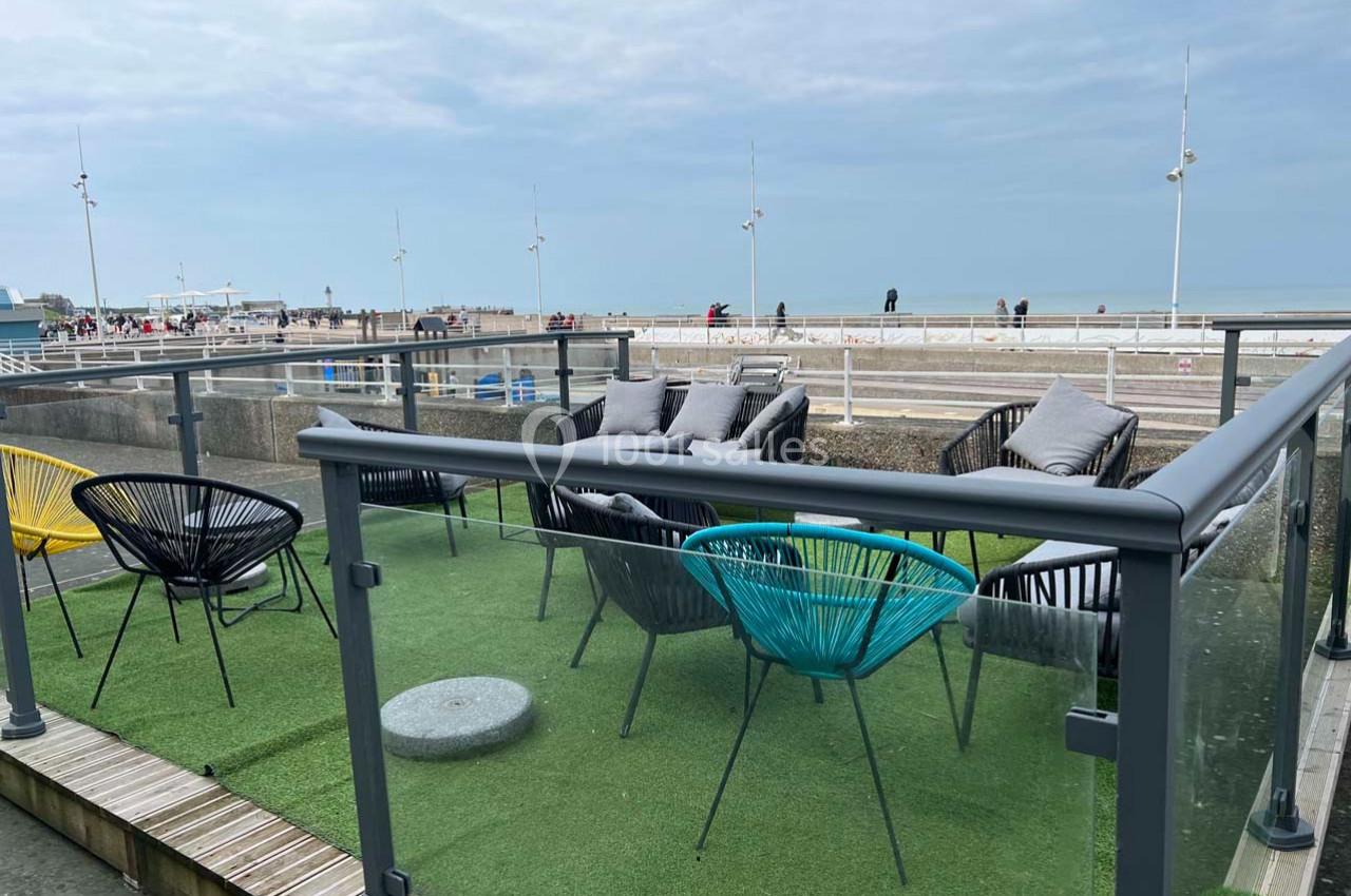 Terrasse avec fauteuils et table basse, vue sur une plage et la mer, entourée d'une balustrade en verre.