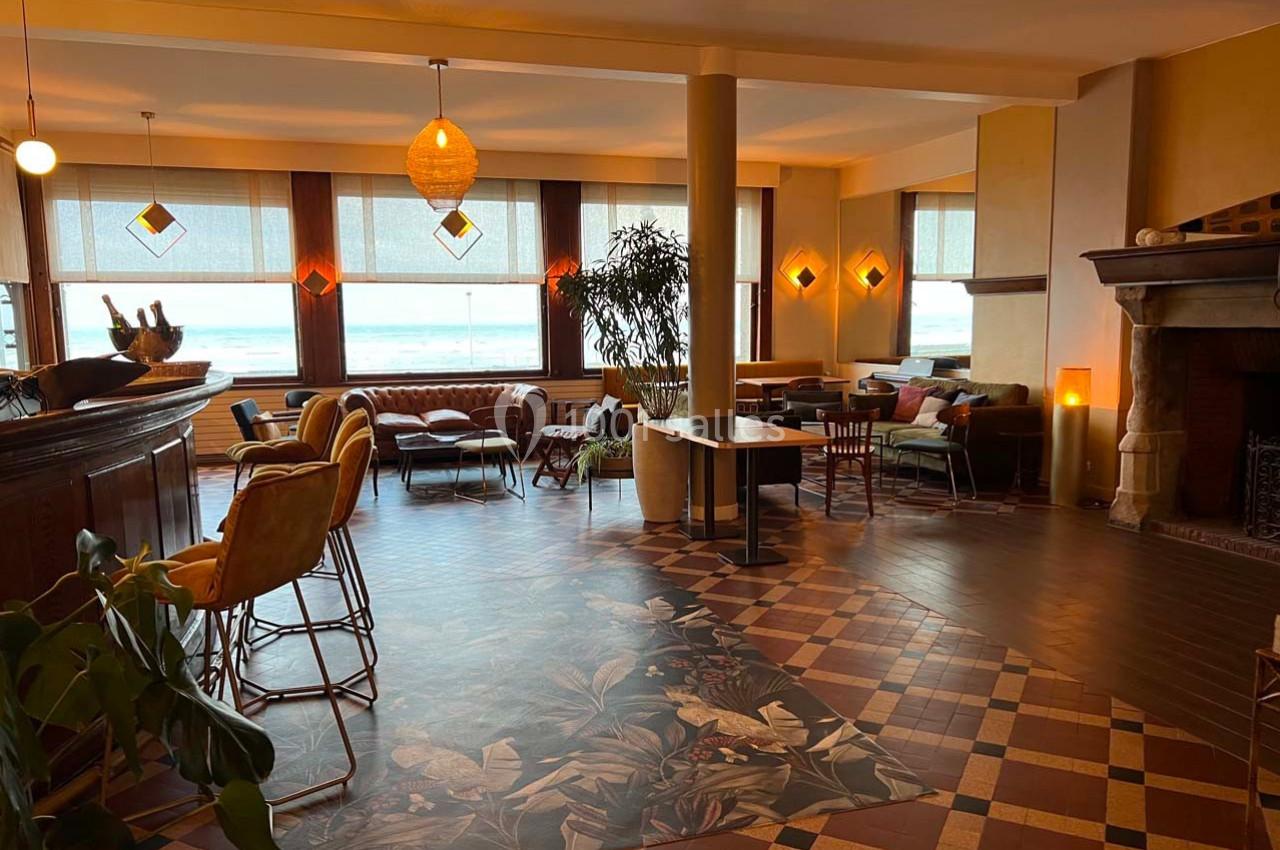 Salle lumineuse avec grandes fenêtres, mobilier varié, sol en carrelage décoratif et cheminée en pierre.