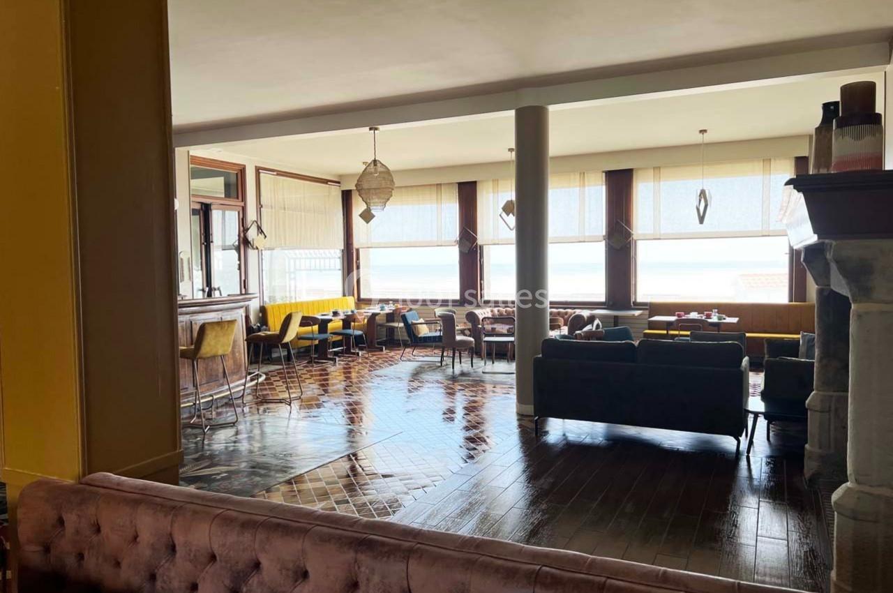 Salle lumineuse avec de grandes fenêtres, mobilier en bois et canapés, offrant une vue sur l'extérieur.