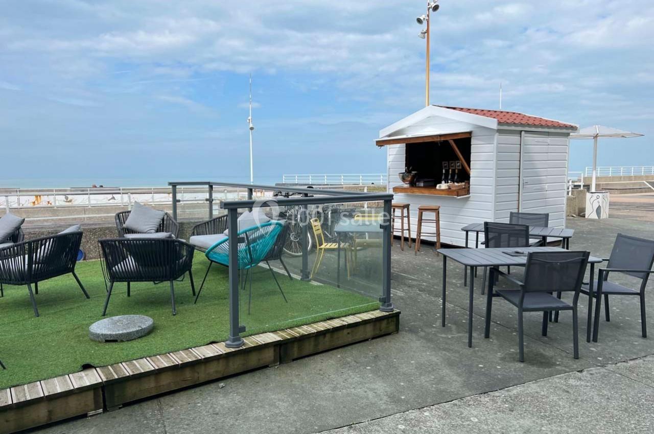 Terrasse en bord de mer avec chaises, tables, espace gazonné et un kiosque blanc sous un ciel partiellement nuageux.
