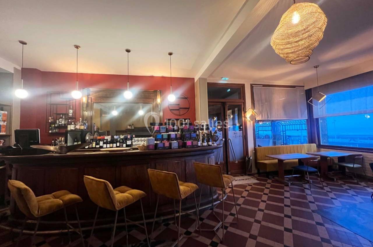 Intérieur d'un bar avec comptoir en bois, tabourets jaunes, tables, banquettes et éclairage suspendu chaleureux.