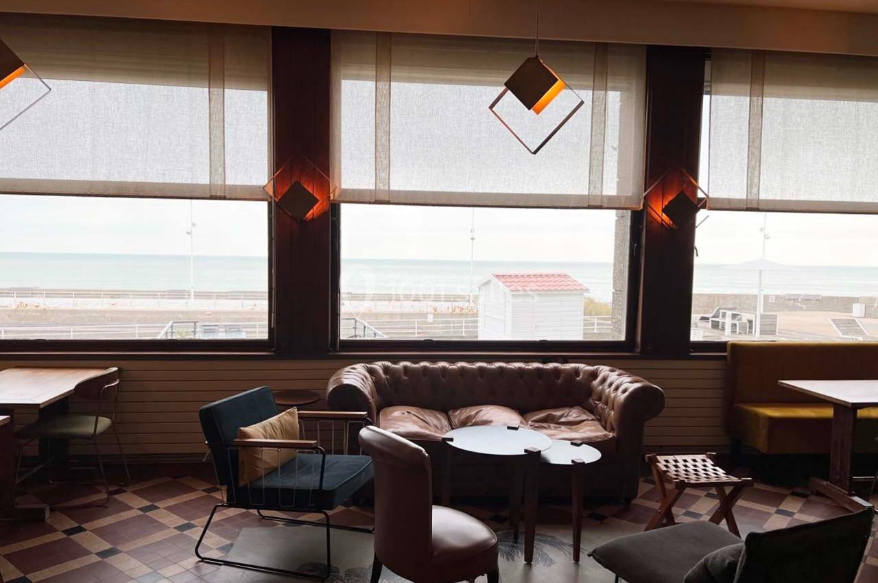 Salon avec fauteuils et tables, grandes fenêtres donnant sur une plage et la mer en arrière-plan.