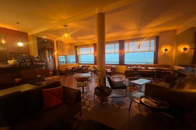 Terrasse en bord de mer avec chaises, tables, espace gazonné et un kiosque blanc sous un ciel partiellement nuageux.