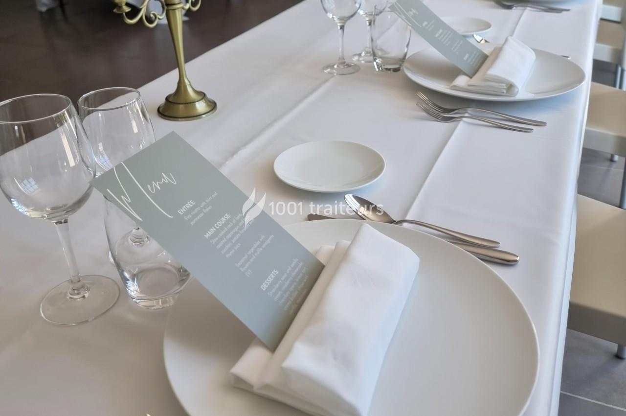 Table dressée avec nappes blanches, assiettes, serviettes pliées, verres, couverts et menus posés sur les assiettes.