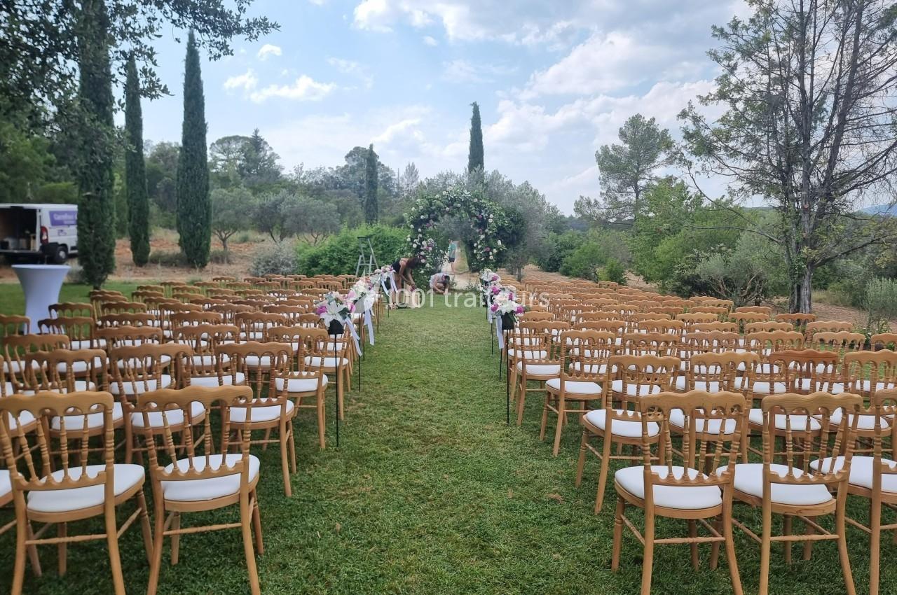 Allée centrale bordée de chaises en bois et décorée de fleurs, menant à une arche florale dans un jardin.
