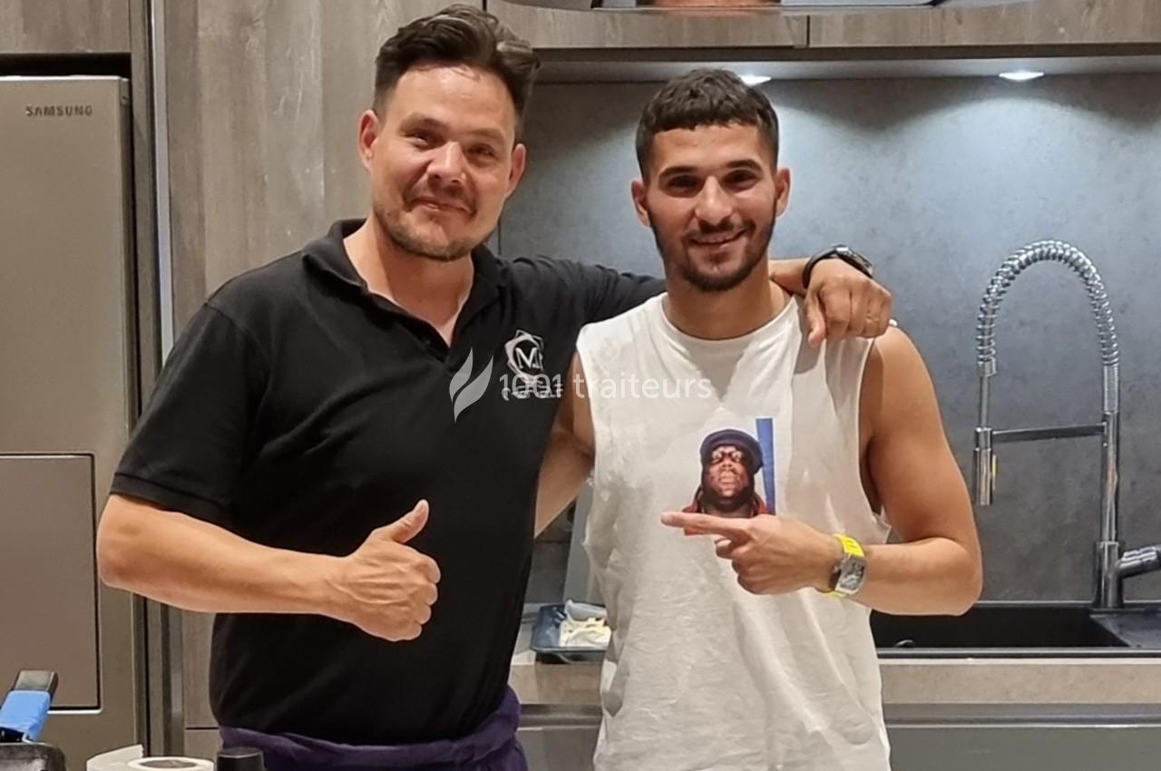 Deux hommes posent dans une cuisine moderne, l'un portant un polo noir et l'autre un débardeur blanc.