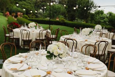 Tables rondes élégamment dressées avec nappes blanches, chaises en bois et guirlandes lumineuses dans un jardin verdoyant.