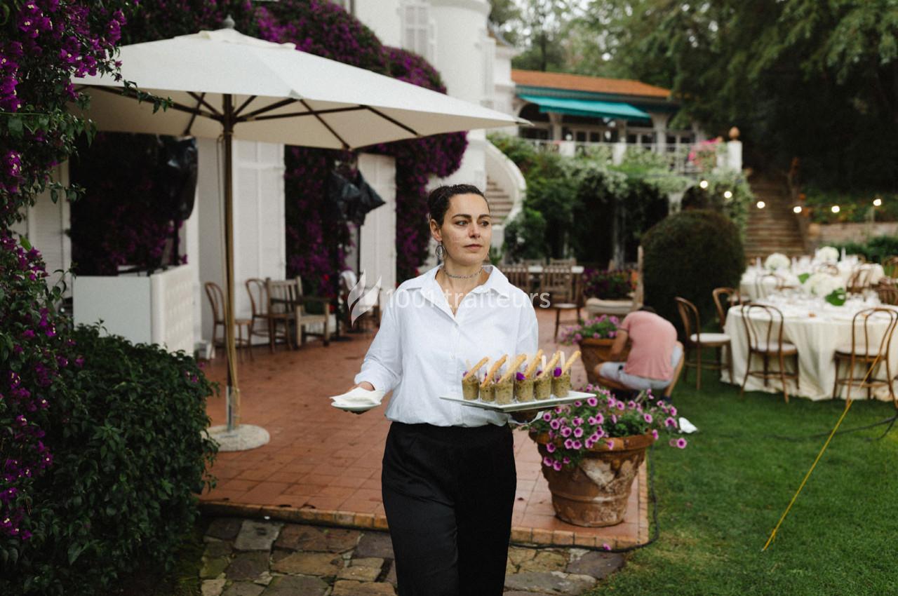 Une serveuse portant des verrines sur un plateau dans un jardin aménagé pour un événement en plein air.