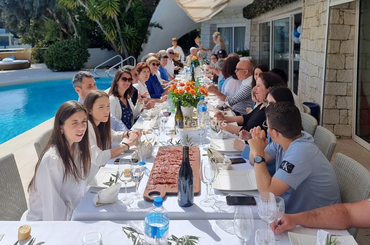 Groupe de personnes assises autour d'une longue table dressée en extérieur, près d'une piscine, partageant un repas.